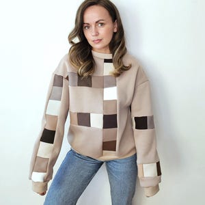 Peut inclure: Un sweat-shirt beige avec un motif patchwork composé de carrés de différentes couleurs neutres, dont le blanc, le marron et le noir. Les manches et le devant du sweat-shirt présentent le motif géométrique. La personne porte un jean bleu.