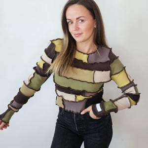 May include: A patchwork long-sleeve top in shades of brown, green, and beige. The top features a ribbed texture and a unique design with contrasting fabric patches. The sleeves are long and fitted, and the top is paired with black jeans.