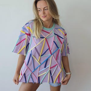 Op de afbeelding: Een lavendelkleurig T-shirt met korte mouwen en een ronde hals met een lichtblauwe bies. De voorzijde en de mouwen zijn versierd met talloze dunne, kleurrijke stoffen stroken in tinten geel, roze, donkerblauw en lichtblauw in een willekeurig patroon.