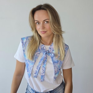 May include: A detachable collar featuring a blue and purple paisley pattern with long fabric ties at the center front. The collar is worn over a plain white short-sleeved t-shirt and blue denim shorts, creating a layered look with a pointed shape.