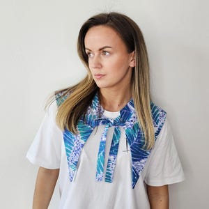 Może przedstawiać: Niebiesko-zielony kołnierz we wzory z wiązaniem noszony jest na białym t-shircie. Kołnierz ma wzór w kształcie liści. Osoba patrzy w bok.