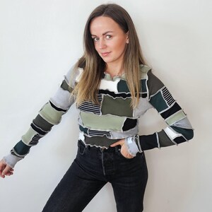 May include: A patchwork long-sleeved top in shades of green, grey, and black. The top features a unique design with various fabric patches. The model is wearing black jeans. The top has a ruffled neckline and cuffs.