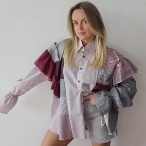 May include: A patchwork button-down shirt in shades of pink, gray, and burgundy. The shirt features a collared neckline, long sleeves with ruffled cuffs, and a mix of patterned fabrics. The model is wearing gray denim shorts.