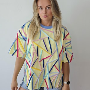 Peut inclure: Un t-shirt à manches courtes jaune pâle doté d'une encolure ronde avec un liseré bleu clair. Le vêtement est orné de nombreuses fines bandes de tissu de différentes couleurs, notamment bleu foncé, rose vif, bleu clair et jaune.