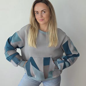 Könnte beinhalten: Ein langärmeliges Sweatshirt mit einem Patchwork-Design in Grau-, Petrol- und Blautönen. Das Kleidungsstück verfügt über einen Rundhalsausschnitt, gerippte Bündchen, einen gerippten Saum und eine große Kängurutasche auf der Vorderseite.