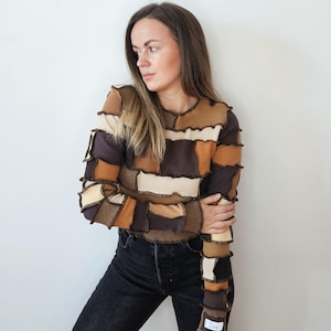 May include: A long-sleeved patchwork top in shades of brown, beige, and cream. The top has a unique design with visible seams and a label on the cuff. The model is wearing black jeans.