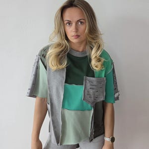 Op de afbeelding: Een patchwork t-shirt in groen, grijs en blauwgroen. Het shirt met korte mouwen heeft een zak en distressed details. Het model draagt een grijze short en een wit horloge.