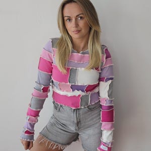 Può includere: Un top a maniche lunghe patchwork nei toni del rosa, viola, grigio e bianco. Il top è realizzato con quadrati di tessuto cuciti insieme con cuciture a vista. La modella indossa pantaloncini di jeans grigi.