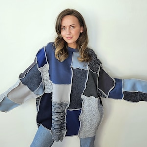 Może przedstawiać: Sweter typu patchwork w odcieniach niebieskiego, szarego i granatowego. Sweter ma różnorodne faktury i kształty tkanin, tworząc unikalny, obszerny design. Modelka ma na sobie niebieskie dżinsy.