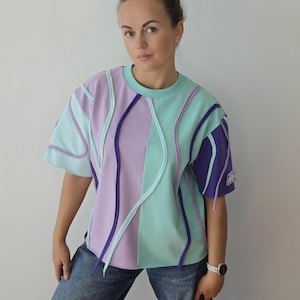 Può includere: Una maglietta a maniche corte con girocollo caratterizzata da un design a blocchi di colore in lavanda, verde menta e viola scuro. Il capo è realizzato con cuciture a rilievo in colori a contrasto che creano un motivo dinamico e ondulato.
