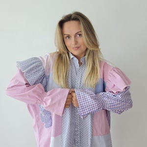 May include: A long-sleeve button-down shirt featuring a patchwork design of various fabrics in shades of pink, light blue, and white. The garment includes panels of gingham, floral prints, and solid colors with ruffled shoulders and smocked cuffs.
