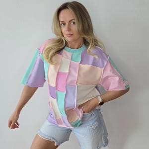 Może przedstawiać: Koszulka patchwork w pastelowych kolorach, w tym różowym, fioletowym i miętowym. Koszulka ma pudełkowy krój i krótkie rękawy. Modelka ma na sobie jasnoniebieskie szorty.