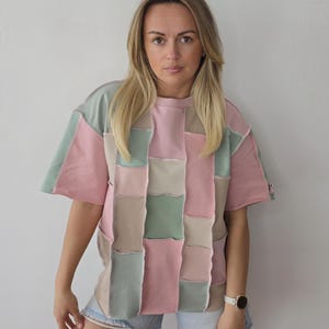 Könnte beinhalten: Ein kurzärmeliges Patchwork-T-Shirt in Pastellfarben, darunter Rosa, Grün und Beige. Das Shirt hat eine kastenförmige Passform und ein einzigartiges, geometrisches Design. Das Model trägt Jeansshorts und eine weiße Uhr.