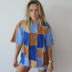 Op de afbeelding: Een t-shirt met korte mouwen voorzien van een patchworkontwerp van rechthoekige stoffen panelen in de kleuren lichtblauw, koningsblauw, beige en bruin. Het kledingstuk heeft een ronde hals en zichtbare naden tussen elk gekleurd stoffen deel.