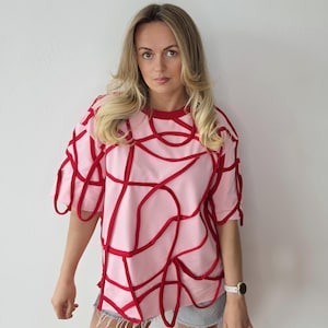 Op de afbeelding: Roze T-shirt met rood abstract ontwerp. De top met korte mouwen heeft een ronde hals en een uniek patroon van rode lijnen over de voorkant en mouwen. Het shirt wordt gecombineerd met een spijkerbroek.