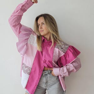 Op de afbeelding: Een overhemd met lange mouwen en knoopsluiting met een patchworkontwerp in verschillende roze tinten, waaronder vichy, effen en bloemmotieven. Het kledingstuk heeft ruches bij de schouders, een kraag en wordt gecombineerd met een grijze spijkerbroek.