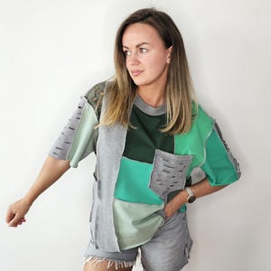 Op de afbeelding: Een patchwork t-shirt in groen- en grijstinten. Het shirt heeft een mix van effen en getextureerde stoffen, met zichtbare stiksels en een zakdetail. Het ontwerp is een unieke, artistieke kijk op een casual t-shirt.