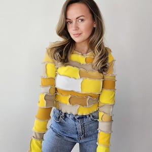 May include: A long-sleeved patchwork top in shades of yellow, beige, and white. The top is paired with blue jeans. The patchwork design features a variety of rectangular and square shapes. The sleeves have a ruffled edge at the wrist.