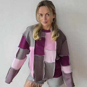 May include: A colour-block sweatshirt in shades of grey, pink, and purple. The long-sleeved top has a crew neck and a unique design with visible seams. The model is wearing grey denim shorts.