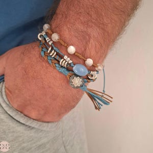 Op de afbeelding: Een gelaagde armband met gevlochten koorden in bruin en blauw, zilveren kralen, witte en blauwe stenen en een zilveren bedel met een stervormig ontwerp.