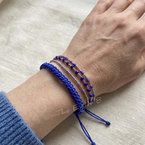 Puede incluir: Una colección de pulseras azules. Una es de diseño trenzado, y las otras son de cuentas azules y doradas. Las pulseras se muestran en una muñeca, mostrando el color y la textura de la joyería. Las pulseras son ajustables.
