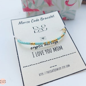 Könnte beinhalten: Ein hellblaues und goldenes Perlenarmband mit einer Karte, auf der "Morse Code Armband", "kryptische Botschaft", "ICH LIEBE DICH MAMA" und eine Website-Adresse stehen.