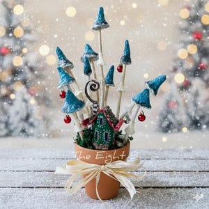 Puede incluir: Un arreglo caprichoso de setas hechas a mano con tapas azules y adornos rojos, exhibido en una maceta de terracota con un lazo decorativo. Una casa verde en miniatura está ubicada entre las setas. El texto "The Eight 8" está en la maceta.