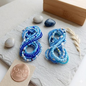May include: Two blue beaded bracelets, each shaped like the number eight. The beads are various shades of blue with silver accents. They sit on a grey stone surface, with a wooden box and decorative stones in the background.