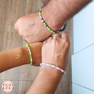 Può includere: Vengono mostrati quattro braccialetti di perline nei toni del verde, blu, giallo, rosa e grigio. La parola "Dad" è stampata su una mano, "Me" su un'altra e "Mom" su una terza. I braccialetti sono indossati sui polsi.