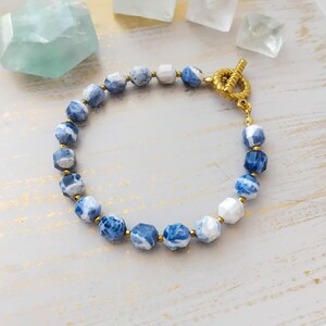 May include: A bracelet made of blue and white faceted beads, interspersed with gold-coloured beads. It has a gold-coloured toggle clasp. The jewellery is displayed on a light-coloured surface with gold accents.