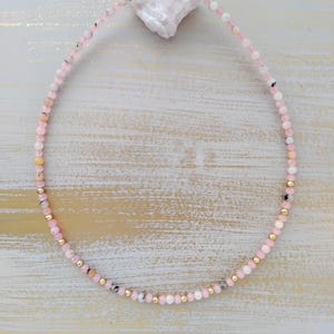 May include: A delicate pink beaded necklace with gold-colored accents. The necklace features small, round beads in shades of pink, white, and gray, interspersed with gold-colored beads. The clasp is also gold-colored.