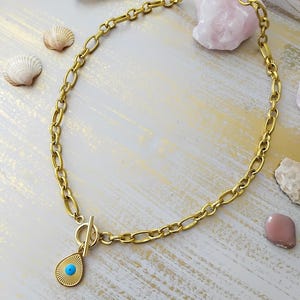 Puede incluir: Collar dorado con una cadena de eslabones grandes y un colgante en forma de lágrima. El colgante presenta un centro azul turquesa y un diseño de rayos de sol. El collar tiene un cierre de palanca. Joyas sobre fondo blanco y dorado.