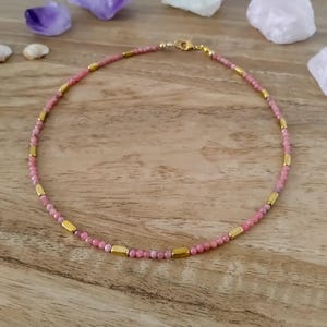 Puede incluir: Un collar de cuentas que presenta pequeñas cuentas redondas de piedra rosa que se alternan con espaciadores metálicos lisos, cilíndricos y de tono dorado. El collar está dispuesto en forma circular e incluye un cierre de mosquetón de metal dorado.