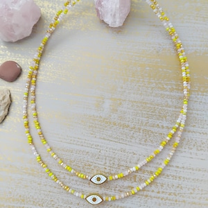 以下が含まれることがあります： 2つの繊細なビーズネックレス。ネックレスは黄色、白、透明のビーズが特徴です。各ネックレスには金色の留め金と、黄色の中心を持つ小さな白い目のチャームが付いています。ネックレスは明るい色のテクスチャード加工された表面に表示されています。
