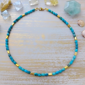 May include: A beaded necklace featuring alternating small turquoise-coloured oval beads and smooth gold-toned cylindrical spacers. The necklace is secured with a gold-toned lobster claw clasp at the top, creating a circular arrangement of blue and gold.
