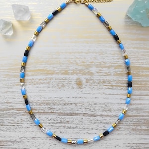 Peut inclure: Un collier de perles composé de perles bleu clair, noires, transparentes et dorées. Le collier est doté d'un fermoir et d'une chaîne dorés. Les perles sont disposées selon un motif symétrique, créant un accessoire délicat et élégant.