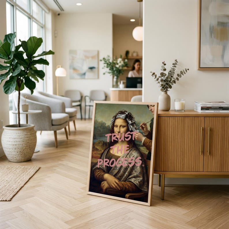 May include: A framed print of the Mona Lisa with the words "TRUST THE PROCESS" in pink graffiti-style lettering. The artwork is leaning against a wooden cabinet in a well-lit room with a plant and chairs.