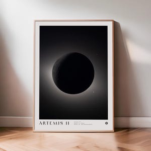 May include: Framed black and white print featuring a celestial body, possibly a planet or moon, with a bright halo effect. The print is labeled "ARTEMIS II" at the bottom, with additional text below. The frame is a light brown color.