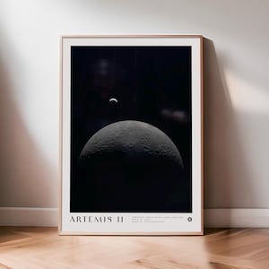May include: A framed print featuring a detailed image of the moon's surface, with a crescent Earth visible in the dark sky. The print is labeled "ARTEMIS II" with additional text below. The frame is a light wood color.