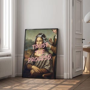 May include: A framed art print of the Mona Lisa, with the words "TRUST THE PROCESS" in pink graffiti-style text. The image is a modern take on the classic painting, with the subject appearing to have her hair styled. The frame is black.