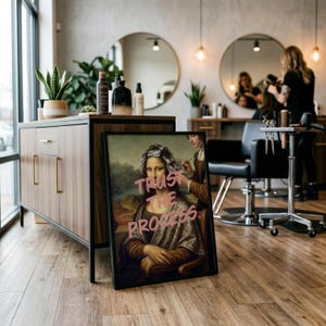 May include: A framed artwork featuring a reimagined Mona Lisa with pink text that reads "Trust the Process." The artwork depicts the Mona Lisa getting her hair styled. The image is in a hair salon setting.