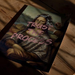 May include: A print of the Mona Lisa with the words "TRUST THE PROCESS" in pink. The artwork is on a wooden surface, with shadows across the image. The classic painting is reimagined with modern text.