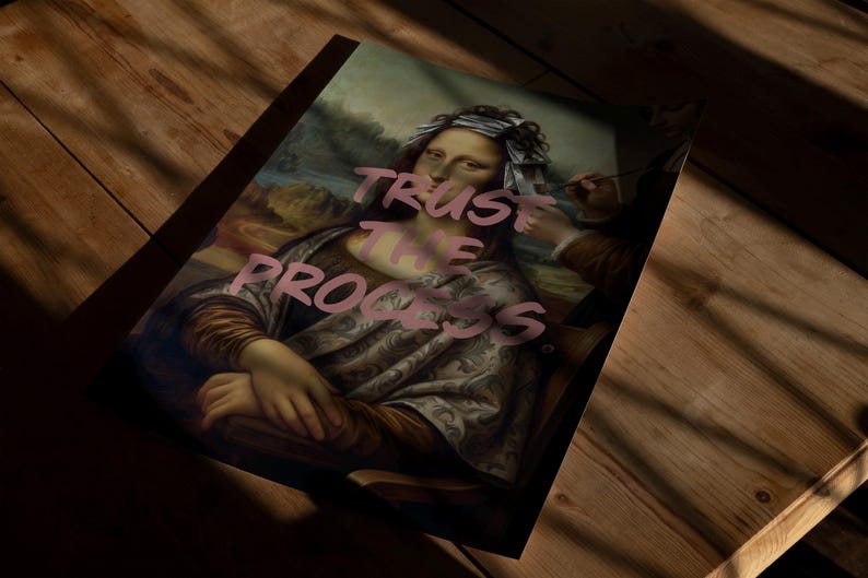 May include: A print of the Mona Lisa with the words "TRUST THE PROCESS" in pink. The artwork is on a wooden surface, with shadows across the image. The classic painting is reimagined with modern text.