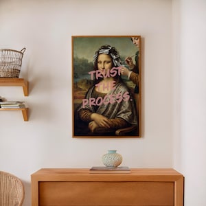 May include: A framed print of the Mona Lisa with the words "TRUST THE PROCESS" in pink graffiti-style lettering. The artwork is in a wooden frame and hangs on a white wall above a wooden cabinet. A small, patterned vase sits on the cabinet.