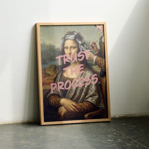 May include: A framed print of the Mona Lisa with the words "TRUST THE PROCESS" in pink graffiti-style lettering. The artwork is in a light wood frame and leans against a white wall. A person is depicted in the background, holding a paintbrush.