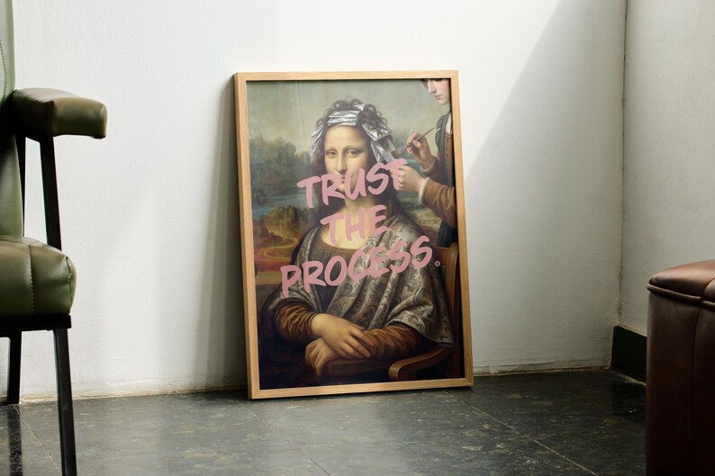 May include: A framed print of the Mona Lisa with the words "TRUST THE PROCESS" in pink graffiti-style lettering. The artwork is in a light wood frame and leans against a white wall. A person is depicted in the background, holding a paintbrush.