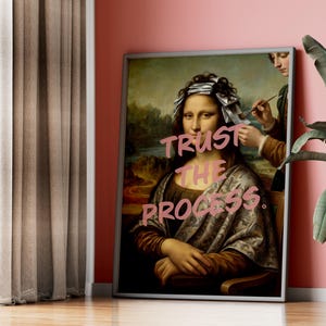 May include: Framed artwork depicting a parody of the Mona Lisa with foil highlights and the text "Trust the Process" in pink. The artwork is displayed against a pink wall, next to a potted plant and a window with brown curtains.