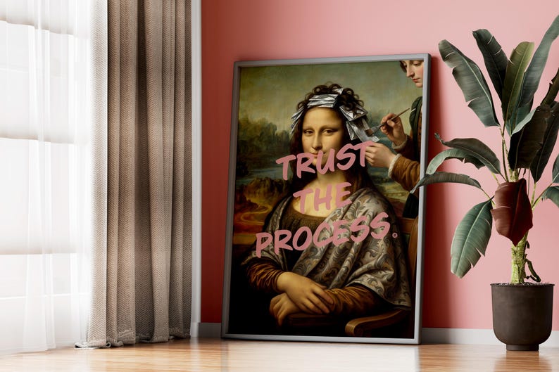 May include: Framed artwork depicting a parody of the Mona Lisa with foil highlights and the text "Trust the Process" in pink. The artwork is displayed against a pink wall, next to a potted plant and a window with brown curtains.