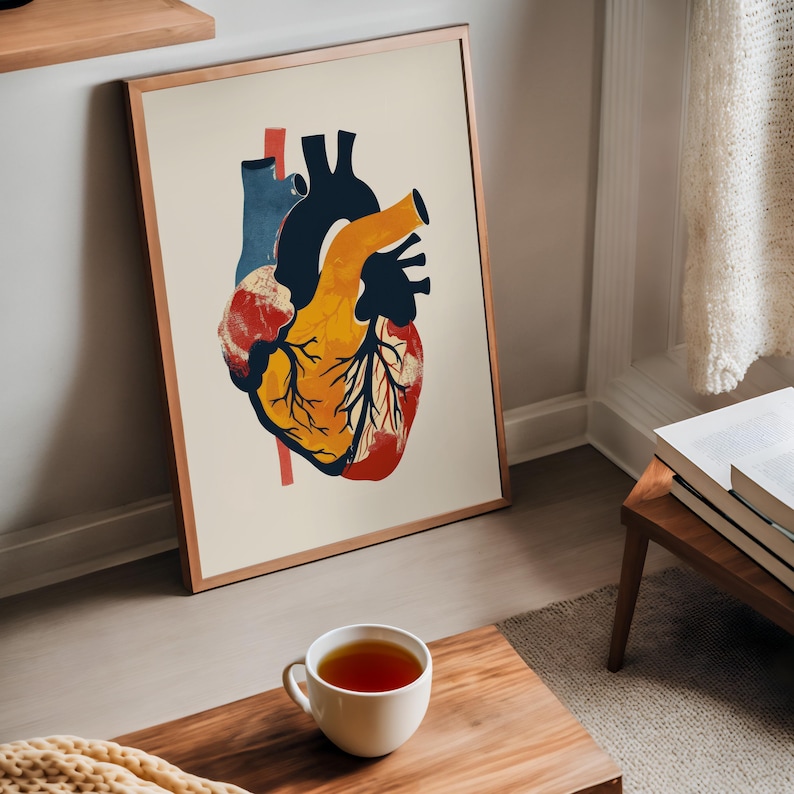 May include: Anatomy-themed art print featuring a stylized human heart illustration. The artwork uses a palette of red, yellow, blue, and black against a cream background. The print is framed in a thin, copper-colored frame and leans against a wall.