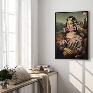 May include: A framed art print of the Mona Lisa with the text "Trust the Process" in pink. The artwork is displayed on a white wall near a window with sheer curtains. The print has a black frame.